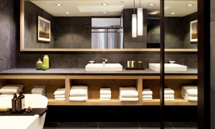 Modern bathroom with a large mirror above a countertop sink, neatly folded towels on wooden shelves, decorative bottles, and a bathtub in the foreground.