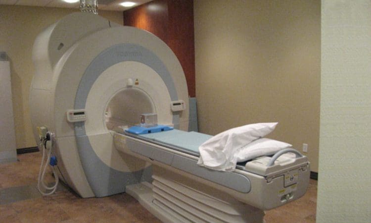 A medical MRI scanner with a patient table and pillow in an exam room with beige walls and a tiled floor.