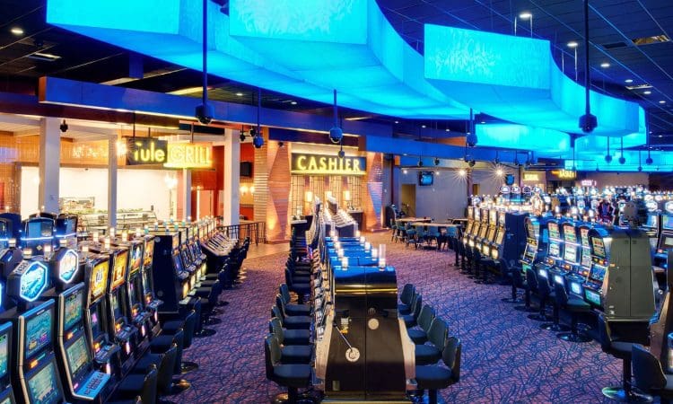 Rows of slot machines in a brightly lit casino, with a glowing blue ceiling feature and a cashier counter in the background.