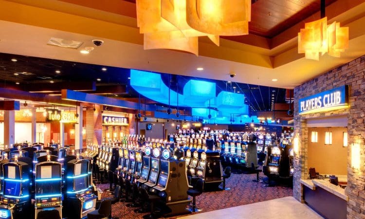 Casino interior with rows of illuminated slot machines, vibrant lighting, and signs for the Tule Grill, Cashier, and Players Club visible.