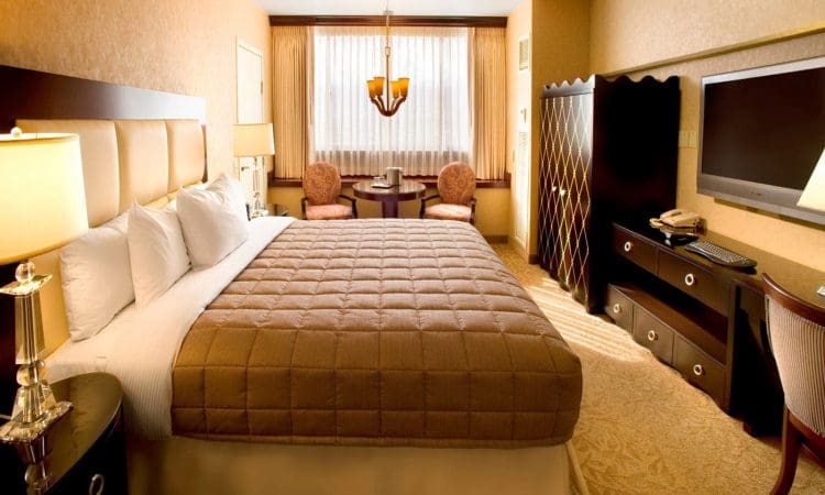 Hotel room with a large bed, bedside tables with lamps, a flat-screen TV on a dresser, a small table with two chairs, and a window with curtains.