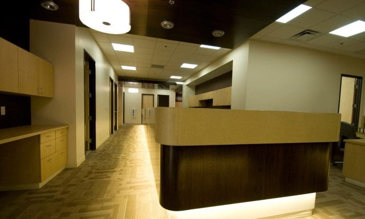 Reception desk in a modern office or medical building with wood and beige finishes, illuminated ceiling lights, and empty hallways.