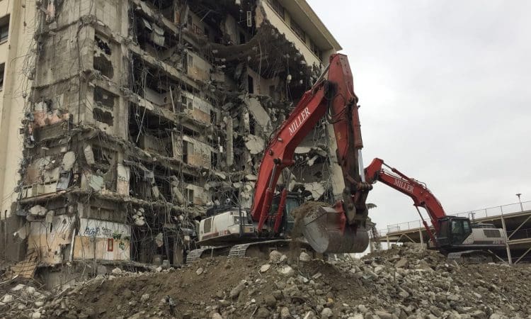 Two excavators demolish a multi-story building, surrounded by rubble and debris under a cloudy sky.