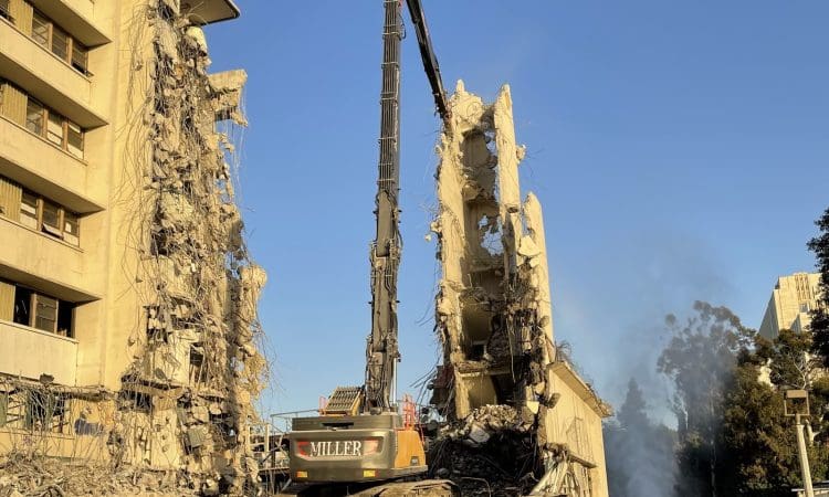 An excavator demolishes a partially collapsed building, with debris and dust rising, under a clear blue sky.