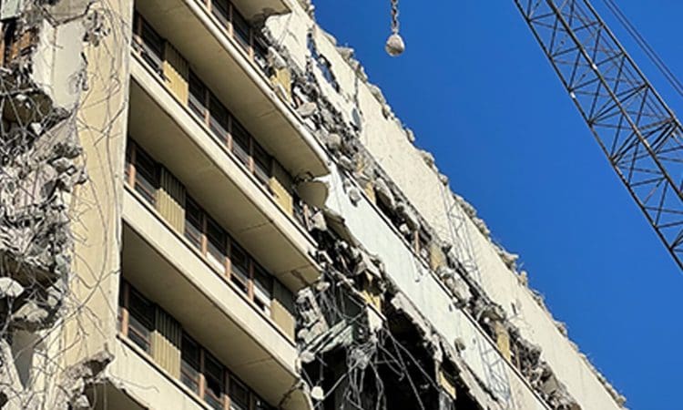 Partially demolished building with exposed floors and debris, a construction crane visible against a clear blue sky.