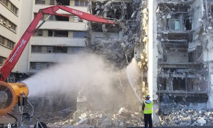 A PENTA construction worker sprays water to control dust while LA County Women's and Children's Hospital is being demolished with heavy machinery. Rubble and debris are visible on the ground.