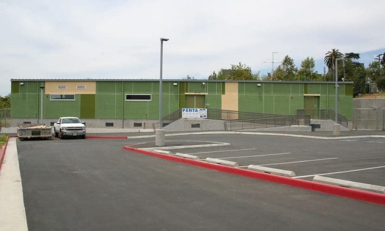 Single-story green modular building with a ramp and empty parking lot in front; a white truck and trailer are parked to the left.