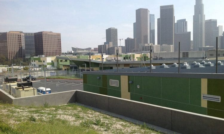 Buildings with green accents and an empty parking lot in the foreground, with the downtown Los Angeles skyline visible in the background on a clear day.