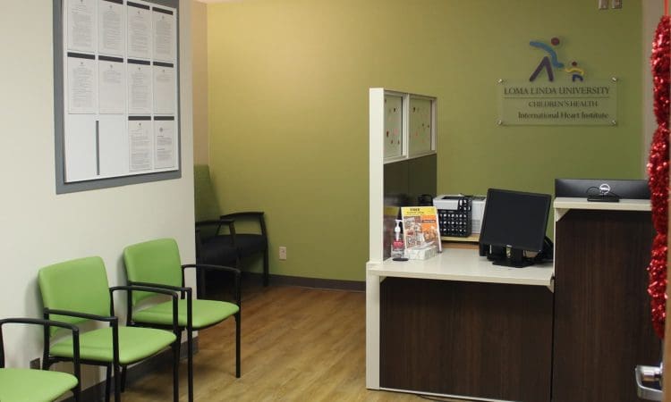 Reception area of a medical office with green chairs, a front desk, a computer, informational brochures, and a wall sign for Loma Linda University Children's Health.