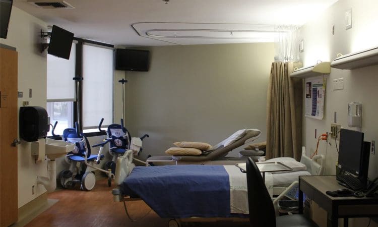 A hospital room with a bed, two chairs, medical equipment, a sink, a computer workstation, and wall-mounted monitors. The room is well-lit with natural and artificial light.