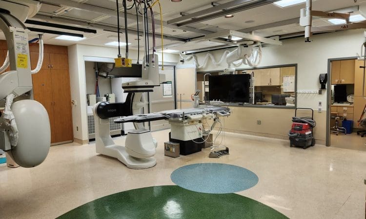 A hospital procedure room with medical imaging equipment, a patient table, monitors, cabinets, and a floor cleaning machine in the corner.