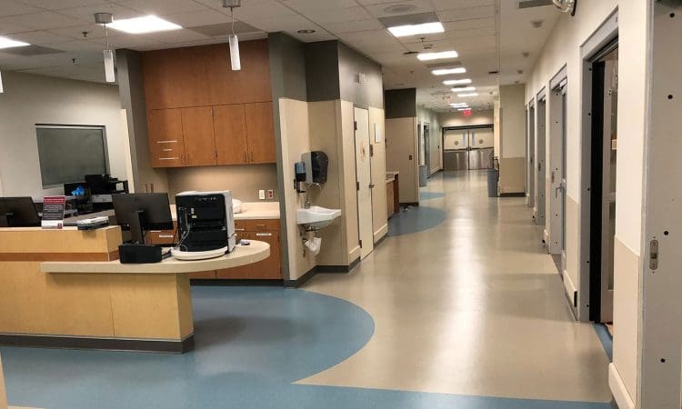 A clean, modern hospital hallway with patient rooms on the right, a nurses’ station on the left, and overhead fluorescent lighting.