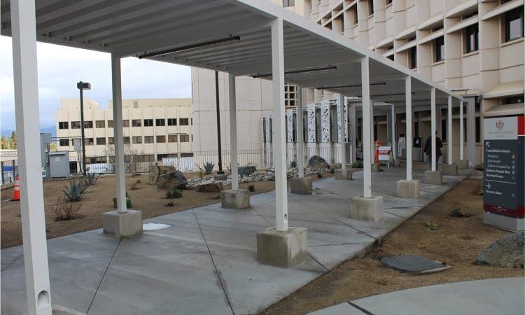 Covered walkway with white metal supports outside a multi-story building, surrounded by dry landscaping and signage.