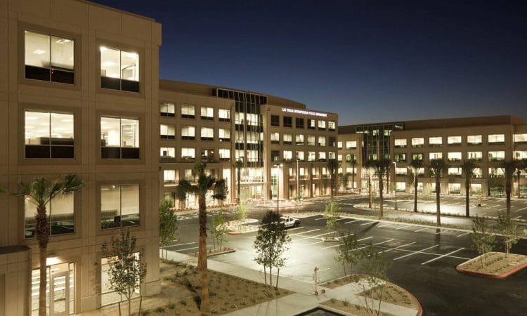 Office complex with multiple lit windows and palm trees in front, surrounding an empty parking lot at dusk.