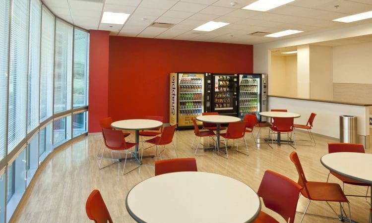 Break room with round tables and red chairs, featuring two vending machines against a red accent wall and large windows along one side.