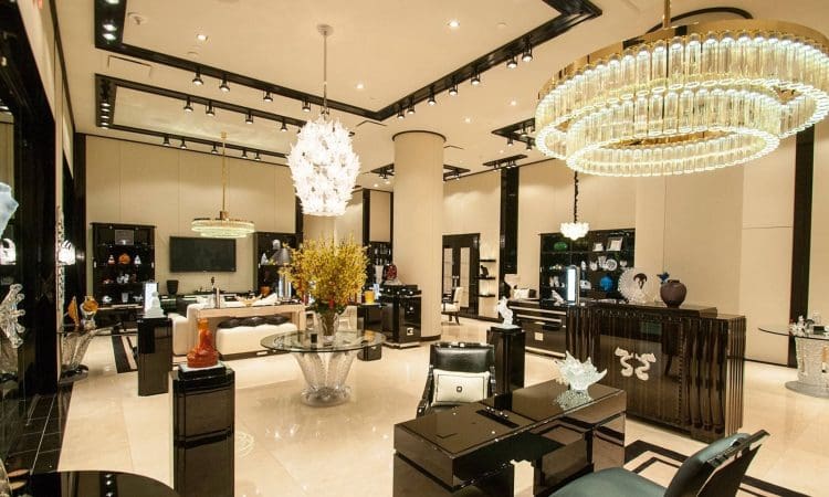 Luxurious, modern showroom with high ceilings, large chandeliers, glossy black furniture, decorative art pieces, and flower arrangements on display tables.