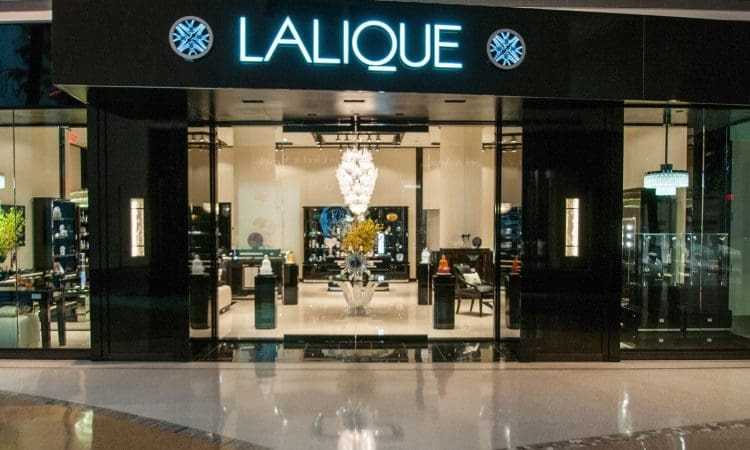 Storefront of a Lalique boutique with a bright neon sign, large glass windows, and a chandelier and decorative items visible inside.