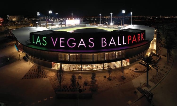 Aerial night view of Las Vegas Ballpark with illuminated signage and stadium lights, surrounded by an empty parking area.