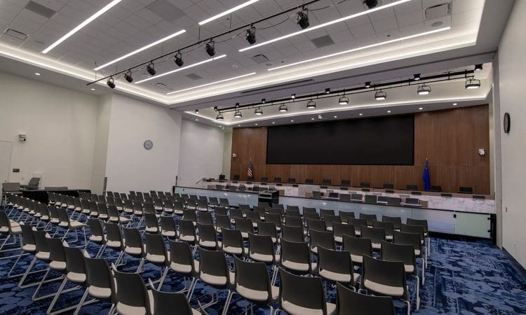 An empty conference room with rows of chairs facing a stage, long desks, flags, and a large screen at the front.