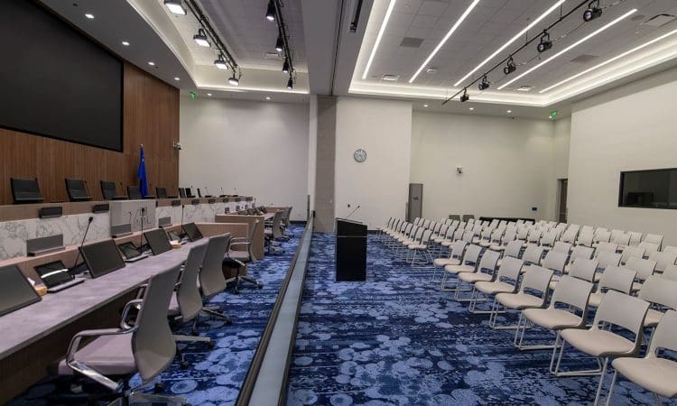 A modern conference room with rows of empty white chairs facing a podium and a long panel desk equipped with monitors and microphones.