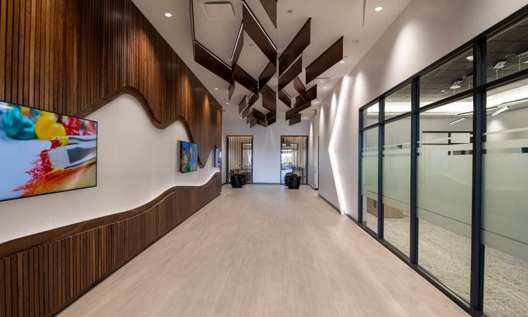 Modern office hallway with wooden wall panels, abstract ceiling design, wall-mounted TVs on the left, and glass-walled offices on the right. The space is well-lit with a light wood floor.