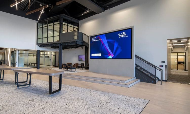 Modern office lobby with a large digital screen on the wall, glass-walled offices, an open staircase, and minimalist tables on a patterned carpet.