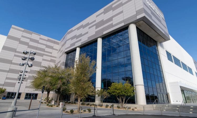 A modern building with large glass windows, tall columns, and a mix of gray and white panels on the exterior, surrounded by trees and a paved area.