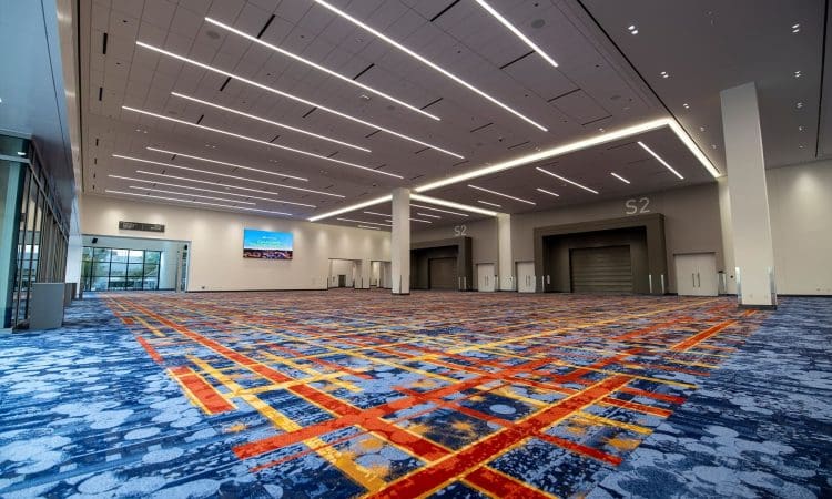 Spacious convention center hall with patterned carpet, high ceilings, bright lighting, and large doors labeled S2. A digital screen displays an image near glass doors.