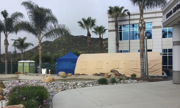 A large tan tent and a blue canopy are set up outside a modern building with palm trees and two portable toilets nearby; mountains are visible in the background.