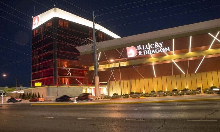 Night view of the Lucky Dragon hotel and casino, with illuminated signs and lights along the exterior facing a street with passing cars.