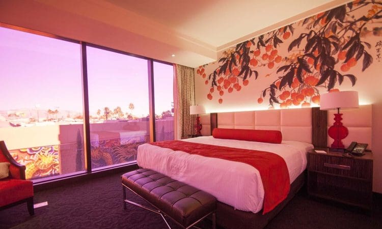 Modern hotel room with a king-sized bed, red and white decor, a floral accent wall, and large windows with a pinkish outdoor view.