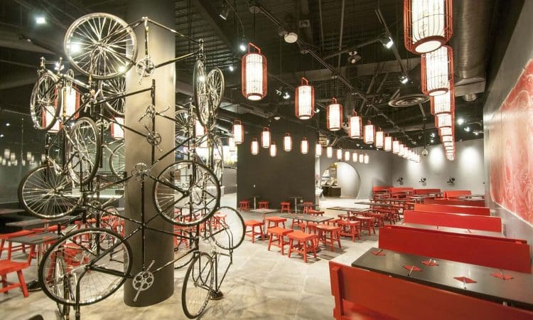 Modern restaurant interior with red tables and chairs, hanging lantern lights, and a decorative column featuring mounted bicycle frames and wheels.