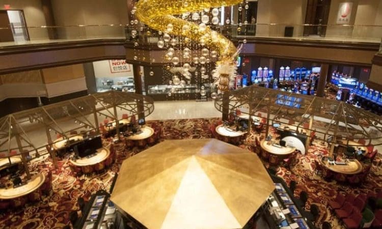 Aerial view of a casino interior featuring a central octagonal cashier area, surrounding gaming tables, slot machines, and a large decorative chandelier overhead.