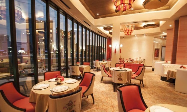 Elegant restaurant interior with round tables, red cushioned chairs, large floor-to-ceiling windows, decorative lighting, and a view of city buildings outside.
