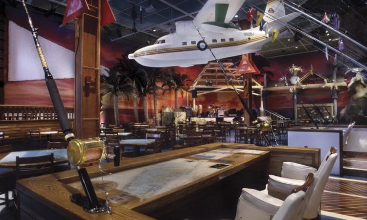 A restaurant interior features nautical decor, a large model seaplane suspended from the ceiling, fishing rods, mounted fish, and wooden tables with chairs.