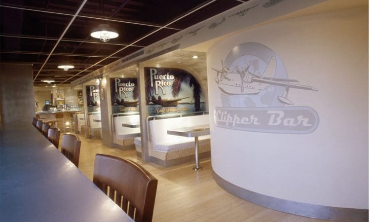 A retro-style diner interior with booth seating, Puerto Rico travel posters, "Clipper Bar" signage, and wooden chairs along a counter.