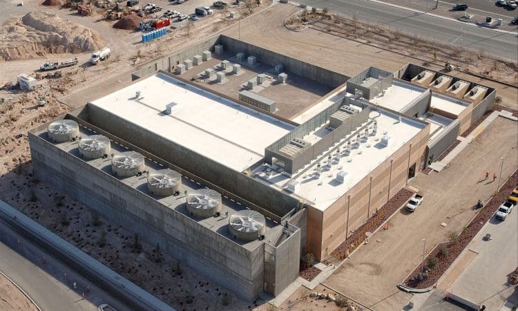 Aerial view of a large, modern data center building with rooftop cooling units, surrounded by desert landscape, parking lots, and nearby roads.