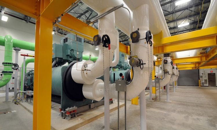 Large industrial pipes and machinery in a facility with yellow support beams, green and white pipes, control panels, and overhead metal ceiling.