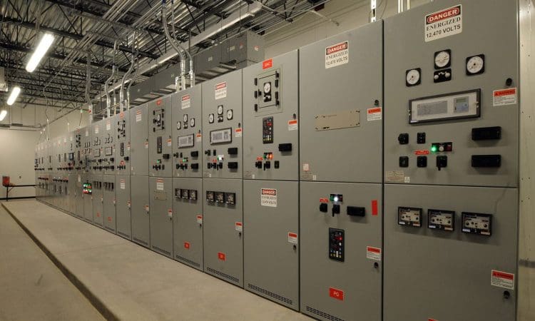 A row of industrial electrical control panels with gauges, switches, and warning labels in a utility room with exposed ceiling and lighting.