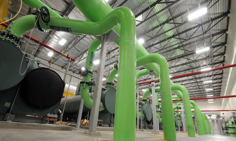 Large green industrial pipes run through a facility with metal grating floors, overhead lighting, and machinery in the background.