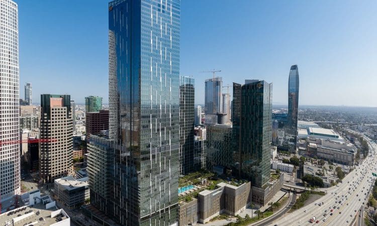 Aerial view of tall glass skyscrapers and high-rise buildings in downtown Los Angeles with a highway in the foreground under a clear blue sky.