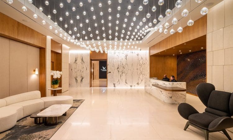 Modern hotel lobby with marble floors, a curved reception desk, lounge seating, decorative lighting fixtures on the ceiling, and two people at the counter.