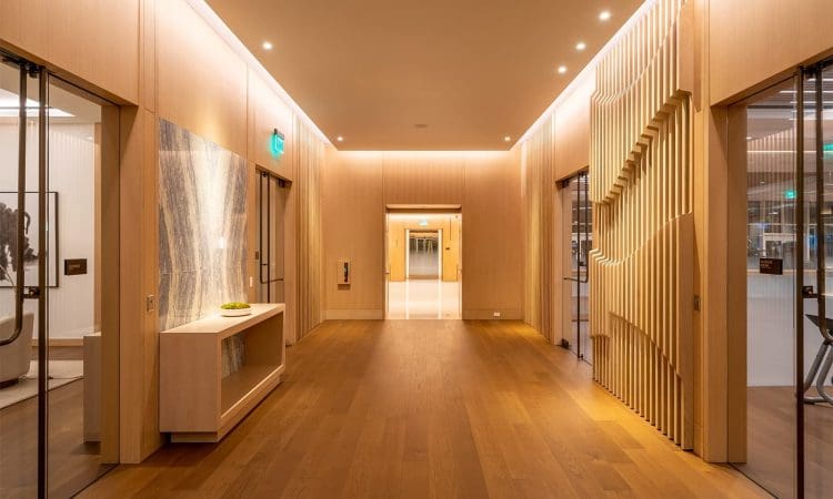 Modern hallway with light wood floors and walls, recessed ceiling lights, decorative paneling, and an elevator at the far end. Glass doors are on both sides.