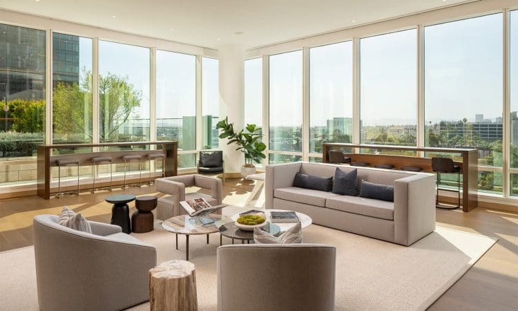 Modern living room with large windows, neutral furnishings, round coffee tables, a wooden stool, and city views. Indoor plant and a long console table add decorative elements.