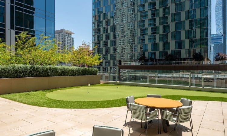 Outdoor patio with round tables and chairs next to a small artificial putting green, surrounded by tall modern glass buildings.