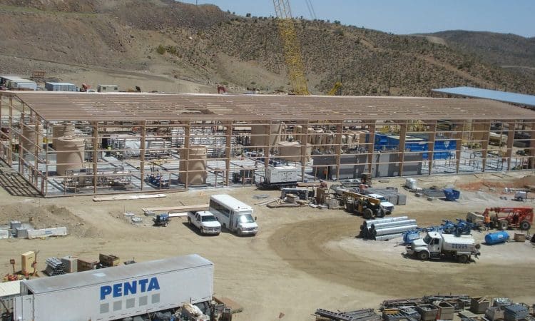 A large industrial building under construction with steel framework, construction vehicles, equipment, and materials spread across a dirt lot at a worksite in a hilly area.