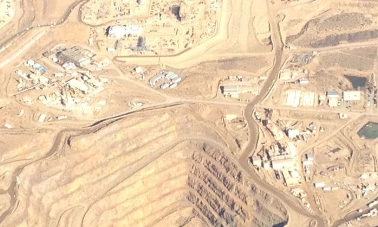 Aerial view of an open-pit mining site with terraced excavation, surrounding buildings, roads, and sparse vegetation in a dry landscape.
