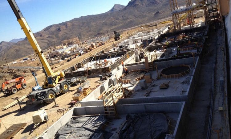 Construction site with workers, cranes, and machinery; concrete structures and building materials are visible with mountains in the background.