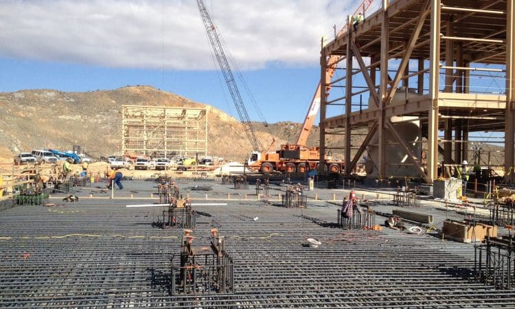 Construction site with steel rebar grid in the foreground, workers, vehicles, a crane, and large steel structures, set against a backdrop of hills and blue sky.