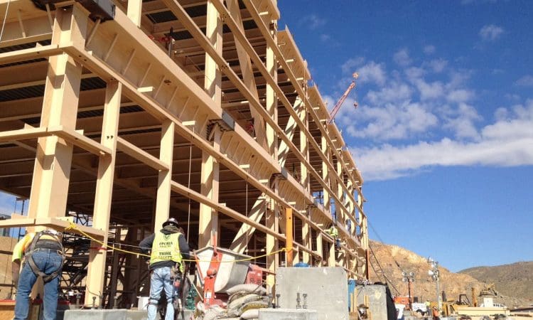 Construction workers in safety gear work on a multi-story building site, with cranes and equipment visible under a clear blue sky.